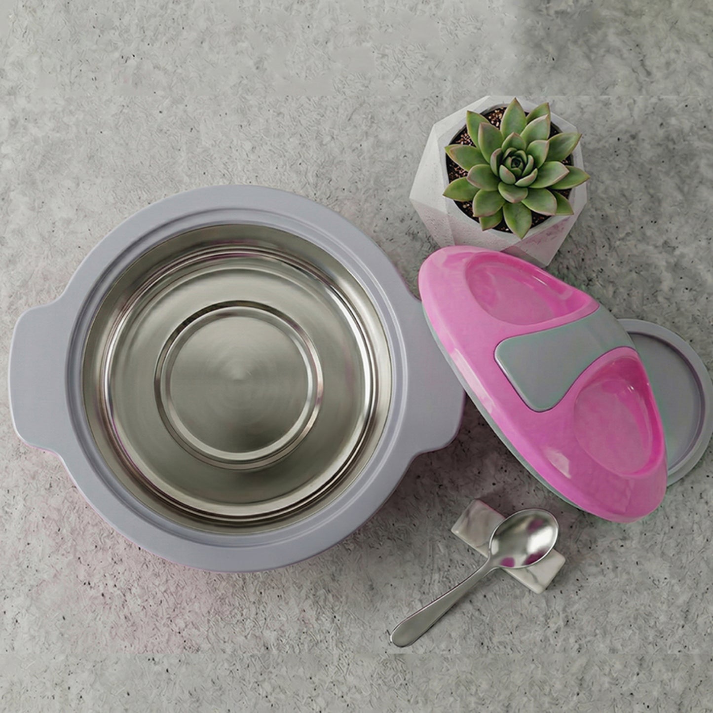 A baby feeding set arranged on a light gray stone surface, including a stainless steel bowl with a light gray silicone liner, a pink and gray silicone spoon holder, a metal spoon, and a small potted succulent.