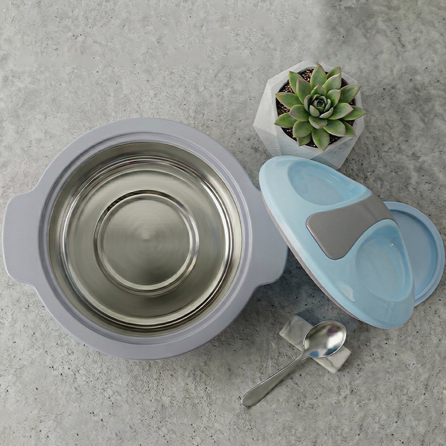 A stainless steel baby feeding bowl with a light purple-gray rim, shown with a light blue and gray divided feeding spoon, a metal spoon, and a small potted succulent on a gray textured surface.